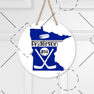 May include: A white round sign with a blue outline of the state of Minnesota. The state is filled in with a solid blue color. The sign has a gray stripe across the middle with the text "Anderson #14" in black. There are two crossed hockey sticks in the middle of the state. A hockey puck is above the text.