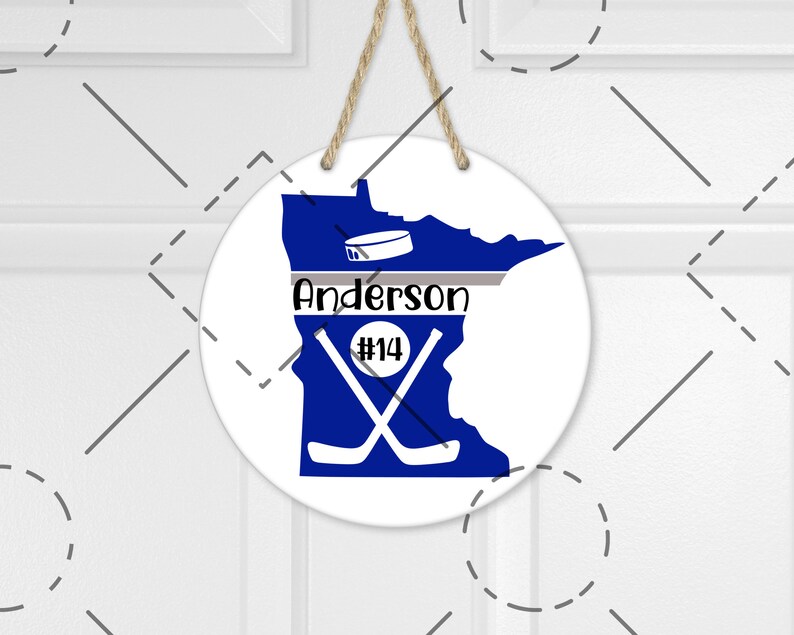 May include: A white round sign with a blue outline of the state of Minnesota. The state is filled in with a solid blue color. The sign has a gray stripe across the middle with the text "Anderson #14" in black. There are two crossed hockey sticks in the middle of the state. A hockey puck is above the text.