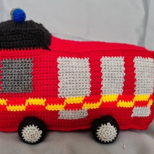 May include: A red crocheted fire truck toy with a black roof, gray windows, and yellow and red stripes. The toy has a blue light on top and gray wheels.