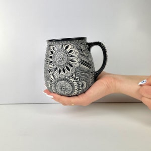 May include: A black and white ceramic mug with a detailed floral and geometric pattern. The mug has a black handle and rim. The design is symmetrical and covers the entire surface of the mug.