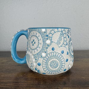 May include: A blue ceramic mug with a white and gray mandala design. The mug has a rounded shape and a handle.