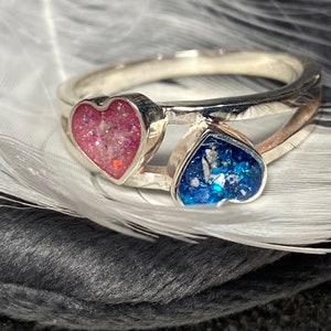 May include: A silver ring with two heart-shaped inlays. The left heart is pink with glitter, and the right heart is blue with glitter.