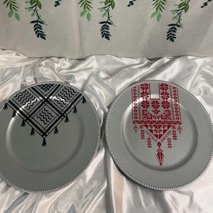 May include: Two silver-colored decorative plates with beaded edges. One plate features a black geometric pattern with tassels, while the other showcases a red floral design. The plates are likely for decorative purposes.