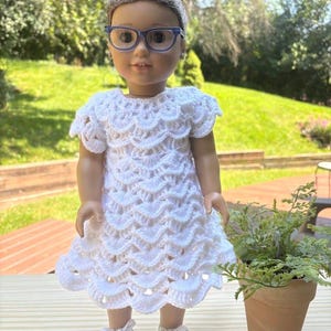 May include: A doll with brown hair styled in a bun, wearing blue-rimmed glasses, a white short-sleeved dress featuring a textured crochet shell pattern, and white lace-up sneakers with silver accents on the toe caps and sides.