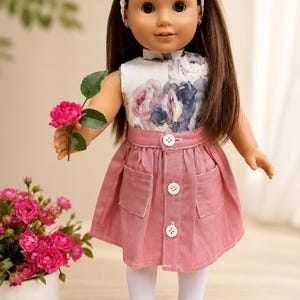 May include: A doll with long brown hair and a white headband, wearing a sleeveless top with a floral print and a pink button-down skirt with two front pockets. The doll has white tights and white slip-on shoes, and is holding a small pink rose.