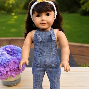 May include: A doll with dark brown hair, wearing a white headband with a small yellow bow, a tan sleeveless top, blue denim-style overalls with cuffed hems, and metallic gold flat shoes. The doll has brown eyes and a neutral facial expression.