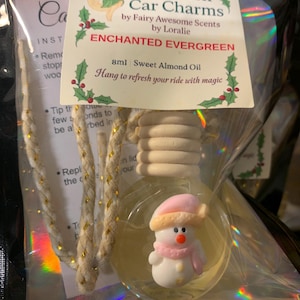 May include: A clear glass car air freshener with a snowman figurine inside. The label reads "ENCHANTED EVERGREEN" and "Hang to refresh your ride with magic." The air freshener is packaged in a clear, iridescent bag with a gold and white rope.