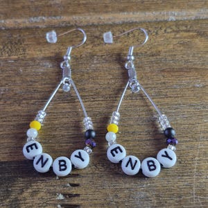 May include: A pair of teardrop-shaped earrings featuring silver-toned wire frames. Each earring is adorned with a sequence of white, yellow, black, and purple beads, including white letter beads that spell out the word ENBY in black capital letters.