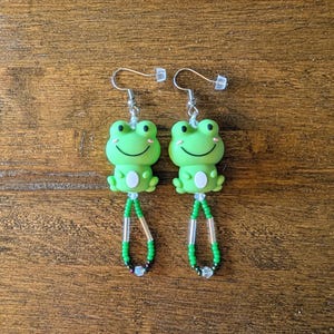 May include: A pair of green frog-shaped earrings with silver hooks. The frogs have white bellies and smiling faces. The earrings dangle with green and black beaded accents.