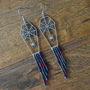 Könnte beinhalten: Ein Paar silberfarbene Halloween-Ohrringe. Jeder Ohrring hat eine Sargform mit einem Spinnennetz-Design und einer kleinen Spinne. Die Ohrringe haben einen Perlenbesatz in Blau-, Grün- und Rottönen, was ein gruseliges Accessoire ergibt.