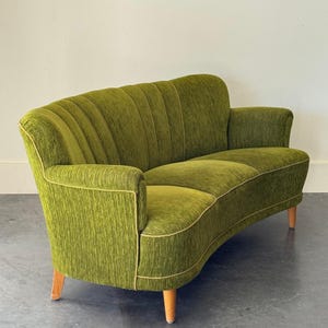 May include: A vintage, curved sofa in a vibrant olive green hue. The sofa features a textured fabric with vertical channel tufting on the backrest and armrests. The edges are trimmed with a light gold piping, and it stands on four wooden legs.