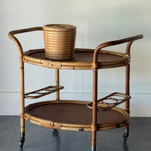 May include: A two-tiered oval rattan serving cart with a light brown finish, featuring four caster wheels, curved handles, and two integrated bottle holders on the lower tier. A cylindrical lidded rattan container sits on the top shelf.