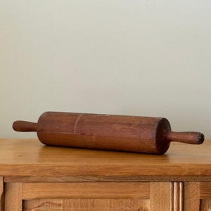 Può includere: Un mattarello vintage in legno con finitura marrone scuro. Il mattarello ha un corpo cilindrico e due manici. È appoggiato su una superficie di legno.