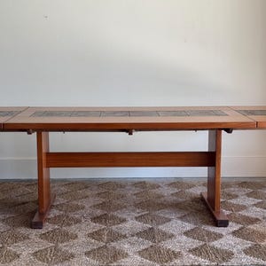 May include: A mid-century modern extendable dining table with a tiled top and a wooden base. The table is made of teak wood and has a natural finish. The table is sitting on a woven rug.