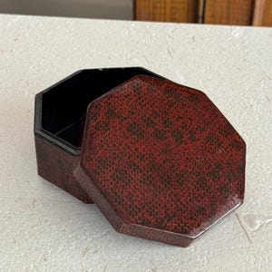 May include: An octagonal box with a removable lid, featuring a dark red and black textured honeycomb pattern on the exterior surfaces. The interior of the box and the underside of the lid are finished in a solid, smooth, glossy black lacquer.