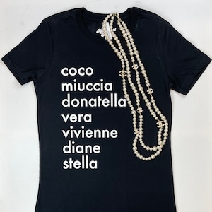 May include: Black t-shirt with white text that reads "coco miuccia donatella vera vivienne diane stella". A pearl necklace with gold charms is draped over the shirt.