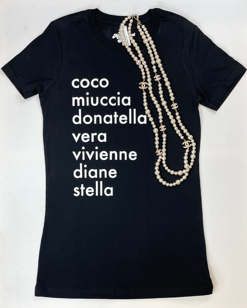 May include: Black t-shirt with white text that reads "coco miuccia donatella vera vivienne diane stella". A pearl necklace with gold charms is draped over the shirt.