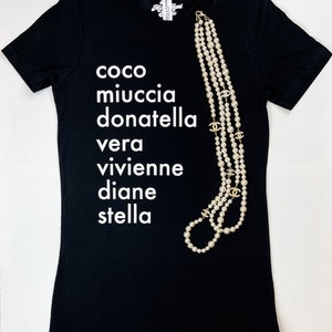 May include: Black t-shirt with white text that reads "coco miuccia donatella vera vivienne diane stella". A pearl necklace with gold accents is draped over the shirt.