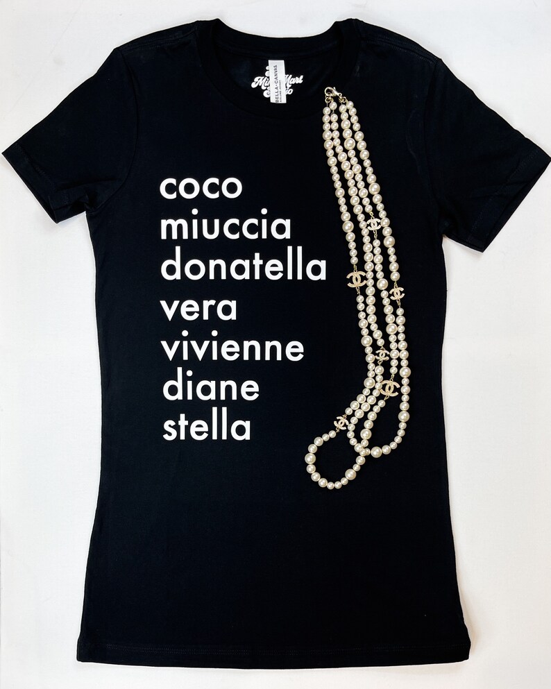 May include: Black t-shirt with white text that reads "coco miuccia donatella vera vivienne diane stella". A pearl necklace with gold accents is draped over the shirt.