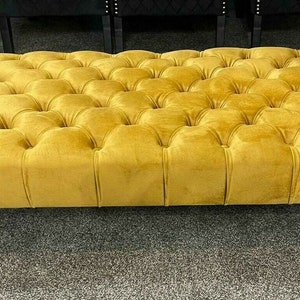 May include: Mustard yellow velvet ottoman with a tufted design. The rectangular ottoman sits on four black, rounded feet. The tufted detailing forms a grid pattern across the surface. The ottoman's dimensions are approximately 150 cm x 90 cm.