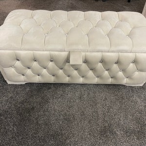 May include: A white tufted storage ottoman with a diamond pattern and crystal-like embellishments. The ottoman has a hinged lid.