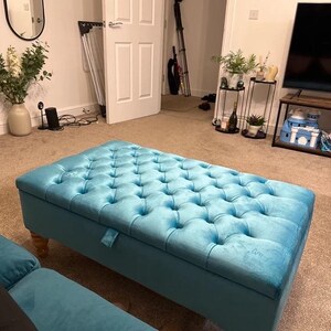 May include: A turquoise blue velvet storage ottoman with a tufted top and wooden legs. The ottoman is sitting on a beige carpet in a living room.