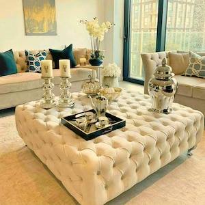 May include: A large, off-white tufted ottoman coffee table with a black tray holding a set of teacups and saucers. The ottoman is decorated with silver candlesticks, a silver vase, and a silver decorative jar.