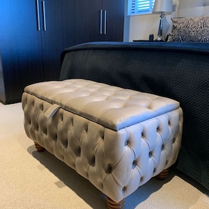 May include: A beige tufted storage ottoman with wooden legs. The ottoman is in a bedroom setting with a bed and a wardrobe in the background.