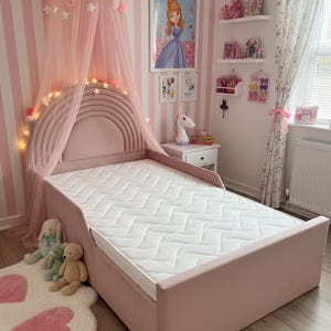 May include: A pink upholstered bed with a rainbow headboard, a white mattress, and a canopy with pink tulle and star lights. The room has pink and white striped wallpaper and decorative shelves.