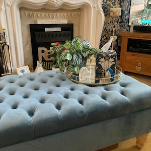 May include: A blue velvet tufted ottoman with wooden legs. The ottoman is in a living room with a fireplace, a plant, and a decorative tray with a tissue box and a sign that says "Love".