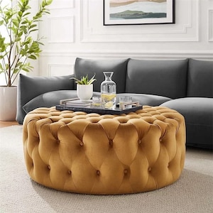May include: A circular, button-tufted ottoman in a mustard yellow hue. It sits before a grey sofa, with a tray holding drinks and a plant. The room's decor features a neutral colour scheme, with a light carpet and white walls.