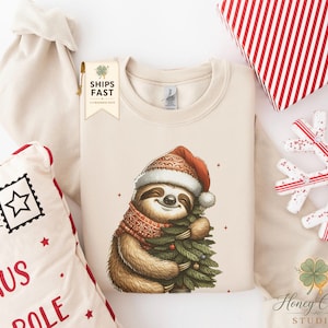 Könnte beinhalten: Ein beigefarbenes Sweatshirt mit Rundhalsausschnitt und einer Grafik eines lächelnden Faultiers, das eine rot-weiße Weihnachtsmütze und einen gemusterten Schal trägt, während es einen kleinen grünen Weihnachtsbaum umarmt. Ein Etikett zeigt SHIPS FAST.