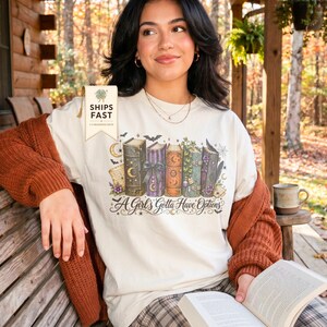 May include: An off-white t-shirt featuring a graphic of five colorful, decorated books with celestial and floral motifs. The text "A Girl's Gotta Have Options" is printed below the books in a decorative script font, accompanied by small stars and moons.
