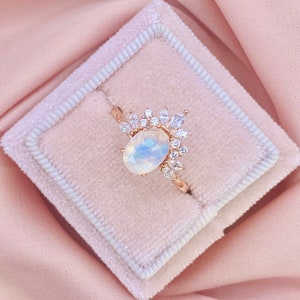 May include: A rose gold ring featuring an oval moonstone centre stone, encircled by smaller, clear accent stones in a sunburst pattern. The ring is presented in a pink velvet ring box.