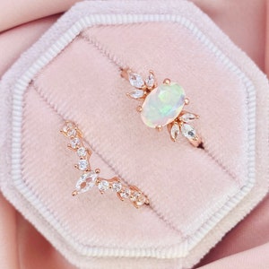 May include: Two rings in a pink velvet ring box. The top ring features a large oval opal surrounded by smaller diamonds. The bottom ring is a delicate band with a row of diamonds.