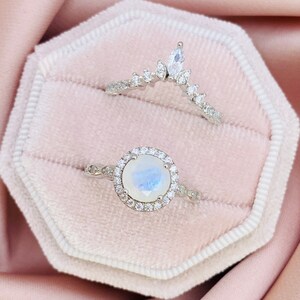 May include: Two silver rings presented in a pink velvet box. One ring has a round, iridescent gemstone encircled by small, clear stones. The second ring is a curved band with a marquise-shaped stone at the top and smaller stones.