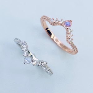 Peut inclure: Deux anneaux délicats avec un design en forme de V, sertis de petites pierres scintillantes et d'une plus grande pierre de lune au centre. L'anneau de gauche est en argent et l'anneau de droite est en or rose.