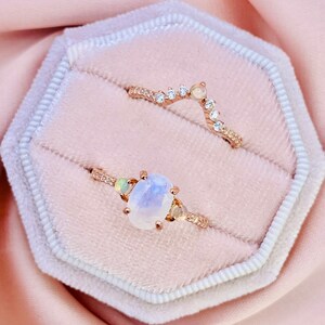 May include: Two gold rings with clear gemstones in a white and pink ring box. The larger ring has a large oval moonstone and two smaller round gemstones. The smaller ring has a curved band with a row of small round gemstones.
