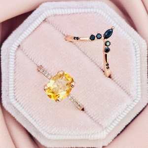Peut inclure: Deux bagues dans un écrin à bijoux en velours. La bague de gauche a une grosse pierre précieuse jaune entourée de petites pierres précieuses claires. La bague de droite a une bande en forme de V avec des pierres précieuses noires.