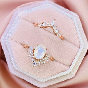 May include: Two rose gold rings set with white gemstones. The larger ring features a large oval-shaped gemstone surrounded by smaller gemstones. The smaller ring is a delicate band with a curved design and smaller gemstones.
