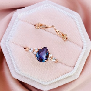 Könnte beinhalten: Ein Paar goldene Ringe in einer weißen Samt-Ringbox. Der Verlobungsring hat einen birnenförmigen, dunkelblauen Edelstein, der von kleineren weißen Edelsteinen umgeben ist. Der Ehering ist ein zartes, gedrehtes Design mit kleinen weißen Edelsteinen.