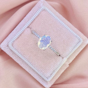 May include: A silver ring featuring a large oval opal gemstone, surrounded by smaller clear gemstones. The ring is presented on a pink velvet ring box.