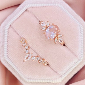 Può includere: Due delicati anelli in oro rosa con pietre di zircone cubico trasparenti. L'anello più grande presenta una pietra centrale di forma ovale circondata da pietre più piccole in un design floreale. L'anello più piccolo è una fascia curva con una pietra centrale di forma marquise e pietre più piccole.