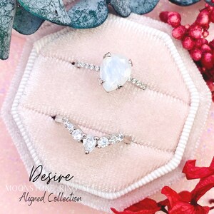 May include: Two silver rings with clear gemstones in a pink velvet ring box. The top ring features a large heart-shaped moonstone. The bottom ring has a curved design with smaller gemstones. The text "Desire Moonstone Ring Set Aligned Collection" is printed below the rings.