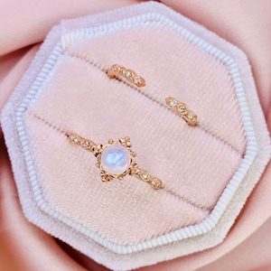 May include: Two gold rings with delicate designs. The larger ring features a round moonstone gemstone surrounded by a floral pattern. The smaller ring has a simple band with small diamonds.