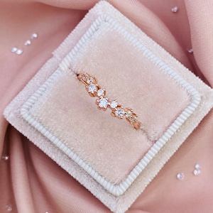 May include: A delicate rose gold ring with a cluster of small diamonds set in a vine-like design. The ring is displayed on a white velvet cushion.