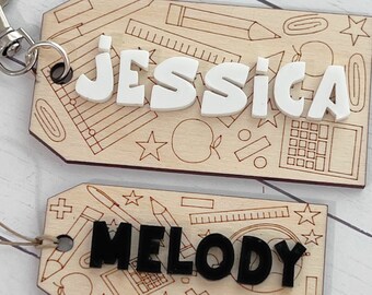 May include: Two wooden tags with a school supply theme. The top tag has the name "JESSICA" in white letters and the bottom tag has the name "MELODY" in black letters.