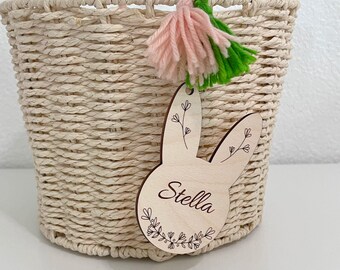 May include: A woven cream-colored basket featuring a light wood tag shaped like a rabbit head. The tag is engraved with the name Stella and floral designs. A pink and green yarn tassel is attached to the top of the tag with a green yarn wrap.