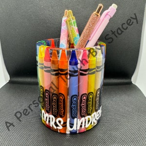 May include: A clear, cylindrical pen holder decorated with colourful crayon designs. The holder is filled with pens of various colours and patterns. The text "MRS UNDRE" is visible on the front. The words "A Personal Touch By Stacey" are also visible.