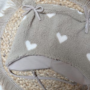 May include: A taupe baby hat with white heart patterns. The hat is made of a soft, textured material and has a chin strap with a bow. It rests on a round, woven mat with a fringed edge. A cosy accessory.